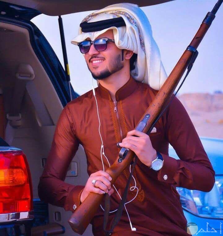 شاب عربي يرتدي عبايه خليجي بني ونظاره وبيده بندقيه
